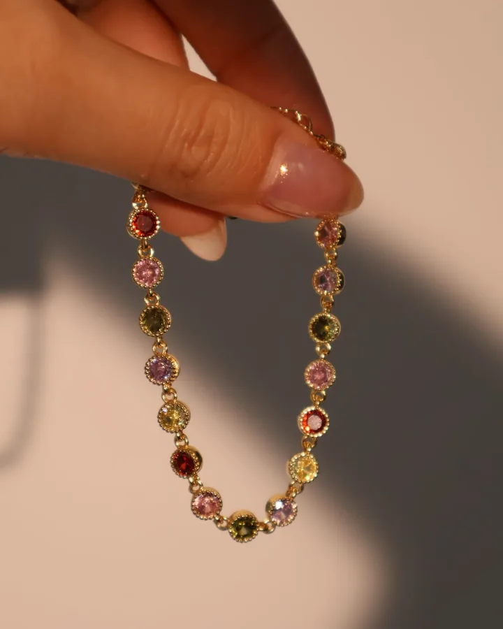 A hand holding a gold bracelet set with pink, red, green and yellow stones